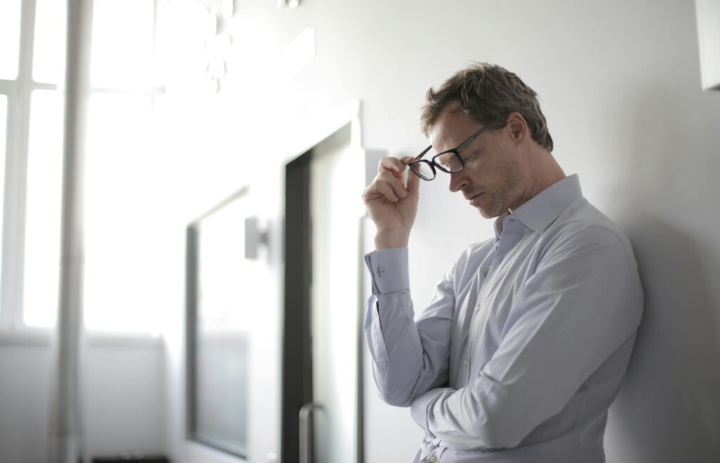 a man standing at a wall holding his glasses in his hands looking down and stressed illustrating neuroticism as Requirements Engineers